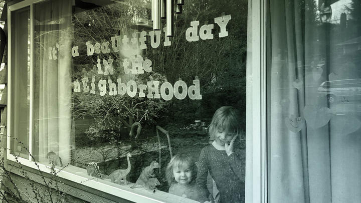 A large window of a house with letters taped up to read &quot;It&#039;s a beautiful day in the neighborhood&quot;, and two children inside